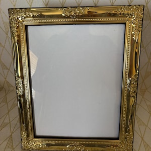 May include: A rectangular picture frame with a polished gold finish and an ornate, embossed floral pattern along the border. The frame features a smooth inner edge and a white rectangular insert, presenting a reflective metallic surface throughout.