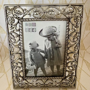 May include: Silver-toned metal photo frame with an ornate, scrolling floral design. The frame holds a black and white photograph of two children. The frame is labeled "SIXTREES" and is designed for a 3" x 5" photo.