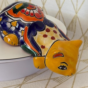 May include: A ceramic cat figurine featuring a vibrant Talavera-style hand-painted pattern. The body displays intricate floral motifs in orange, dark blue, green, and white, while the head is a solid yellow with black painted eyes and nose details.