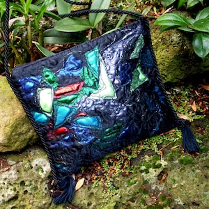 Pode incluir: Uma bolsa acolchoada preta com uma alça longa e um tassel. A bolsa é decorada com formas geométricas coloridas em tons de azul, verde e vermelho.