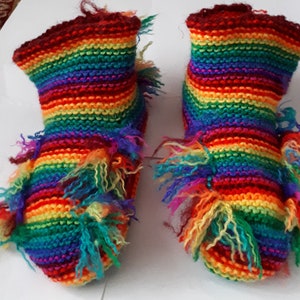 Puede incluir: Un par de zapatillas de punto a rayas de arcoíris con ribete peludo. Las zapatillas están hechas con una variedad de colores, incluyendo rojo, naranja, amarillo, verde, azul, índigo y violeta.