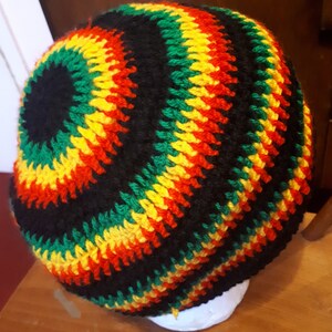 Puede incluir: Un gorro de punto de ganchillo en negro, verde, amarillo y rojo. El gorro tiene un dise&ntilde;o circular con rayas de cada color.