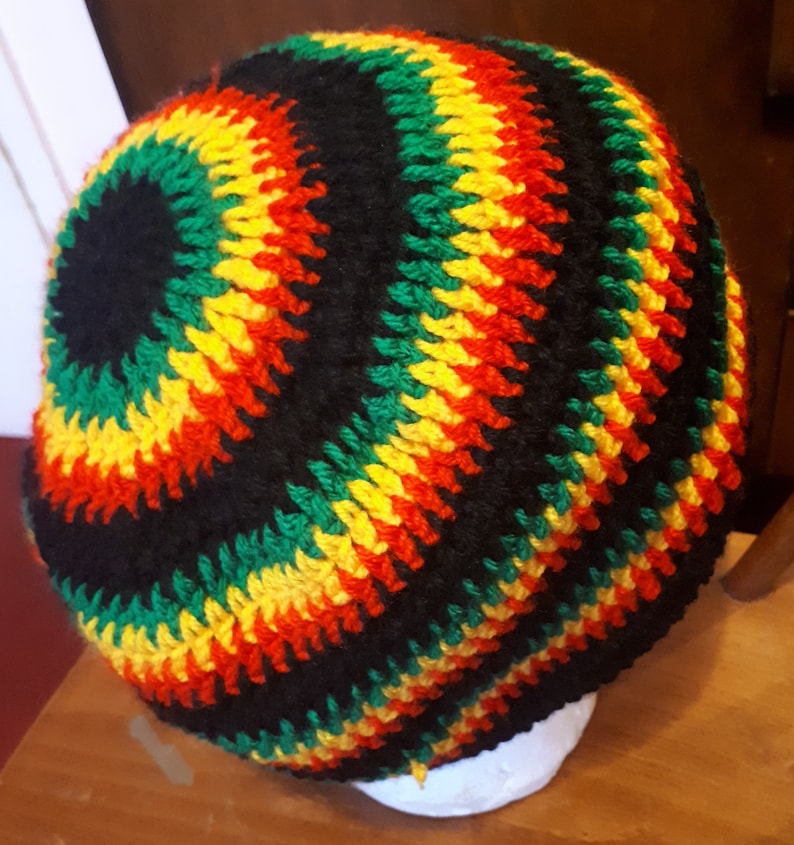 Puede incluir: Un gorro de punto de ganchillo en negro, verde, amarillo y rojo. El gorro tiene un dise&ntilde;o circular con rayas de cada color.