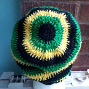 Puede incluir: Un gorro de punto de crochet en negro, verde y amarillo. El gorro est&aacute; hecho con un patr&oacute;n circular y tiene una tela gruesa y texturizada.