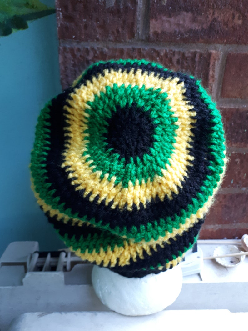 Puede incluir: Un gorro de punto de crochet en negro, verde y amarillo. El gorro est&aacute; hecho con un patr&oacute;n circular y tiene una tela gruesa y texturizada.