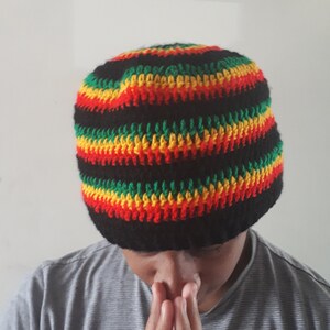 Puede incluir: Un gorro negro de crochet con rayas rojas, amarillas y verdes. El gorro lo lleva puesto una persona con las manos juntas delante de ella.