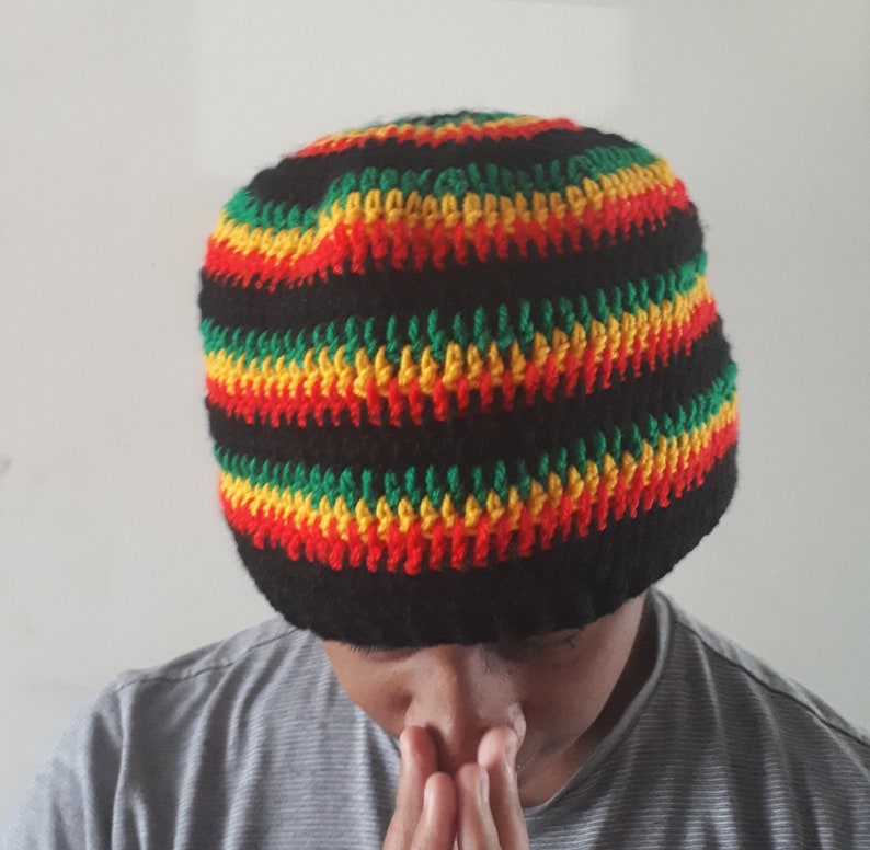Puede incluir: Un gorro negro de crochet con rayas rojas, amarillas y verdes. El gorro lo lleva puesto una persona con las manos juntas delante de ella.