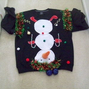 Op de afbeelding: Zwarte kersttrui met een sneeuwmanontwerp. De sneeuwman is gemaakt van wit vilt en heeft een wortelneus, zwarte ogen en een rode mond. De trui is versierd met groene en rode tinsel en heeft een rode pompon bovenop.