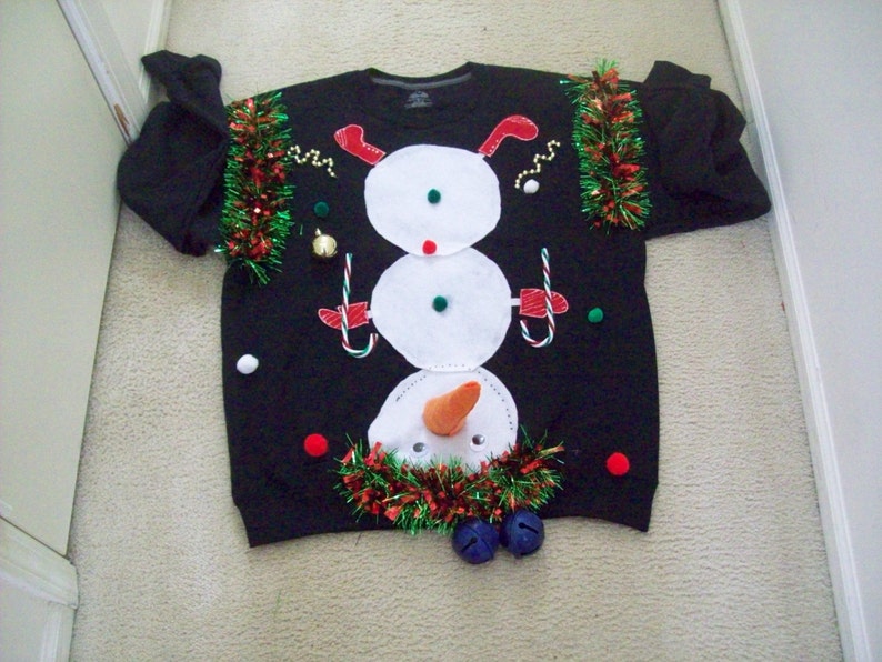 Op de afbeelding: Zwarte kersttrui met een sneeuwmanontwerp. De sneeuwman is gemaakt van wit vilt en heeft een wortelneus, zwarte ogen en een rode mond. De trui is versierd met groene en rode tinsel en heeft een rode pompon bovenop.