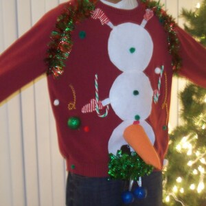 Op de afbeelding: Een rode kersttrui met een sneeuwpopontwerp. De sneeuwpop heeft een wortelneus, groene en witte snoepstokken en een groene krans om zijn nek. De trui is versierd met groene en rode tinsel.