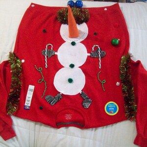 Op de afbeelding: Rode kersttrui met een sneeuwpopdesign. De sneeuwpop is gemaakt van wit vilt en heeft een wortelneus, zwarte ogen en een zwarte hoed. De trui is versierd met groene en rode kerstversiering.