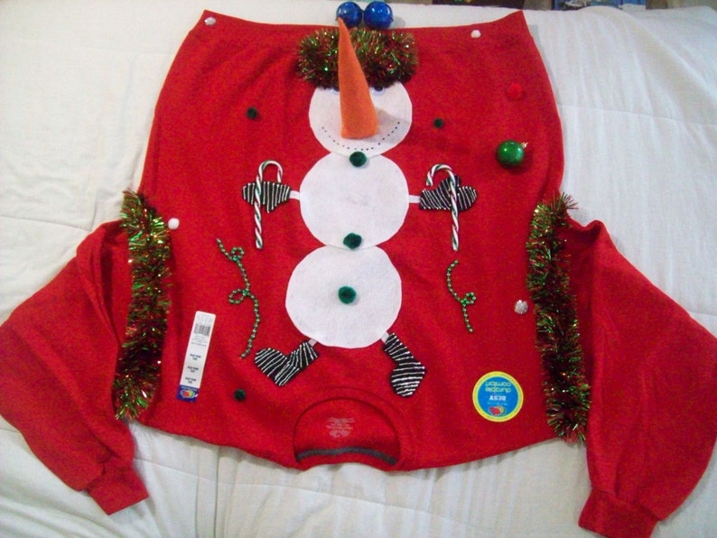 Op de afbeelding: Rode kersttrui met een sneeuwpopdesign. De sneeuwpop is gemaakt van wit vilt en heeft een wortelneus, zwarte ogen en een zwarte hoed. De trui is versierd met groene en rode kerstversiering.