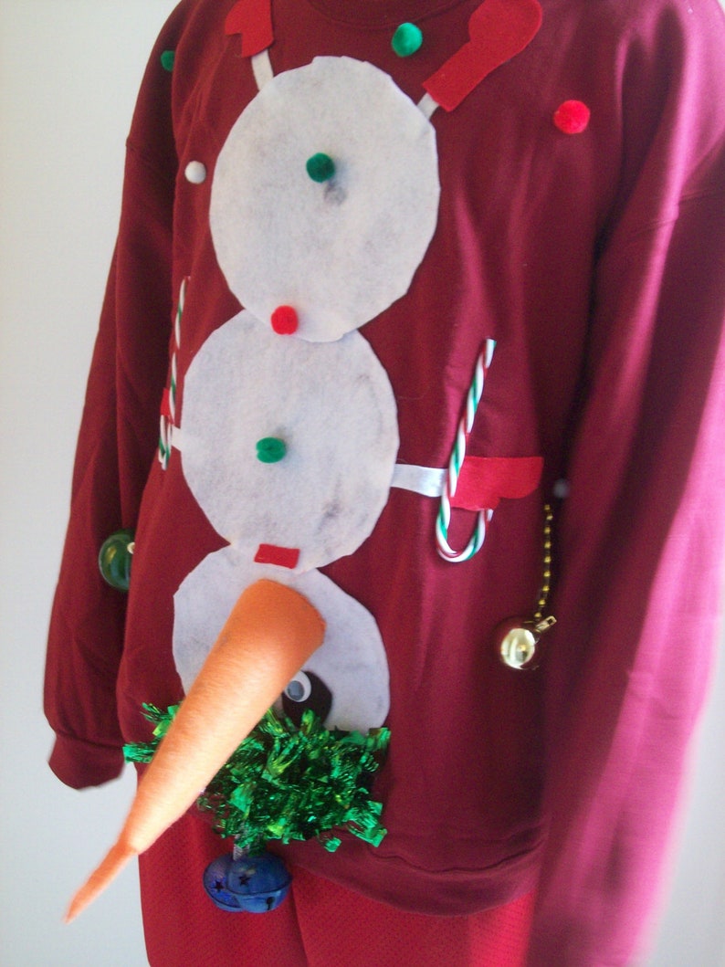 Op de afbeelding: Een rode kersttrui met een sneeuwpopdesign. De sneeuwpop is gemaakt van wit vilt en heeft een wortelneus, groene knopen en een zwarte hoed. De trui heeft ook groene en rode viltversieringen.