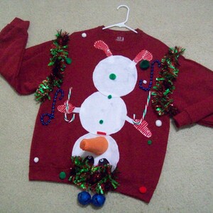Op de afbeelding: Rode kersttrui met een sneeuwpopdesign. De sneeuwpop heeft een wortelneus, zwarte ogen en een wit lichaam. De trui is versierd met groene en rode tinsel en witte kralen.