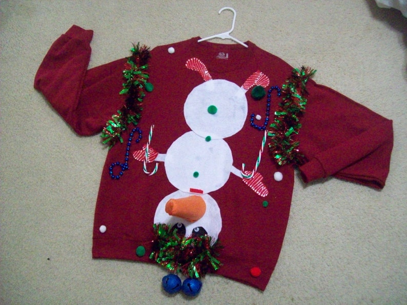 Op de afbeelding: Rode kersttrui met een sneeuwpopdesign. De sneeuwpop heeft een wortelneus, zwarte ogen en een wit lichaam. De trui is versierd met groene en rode tinsel en witte kralen.