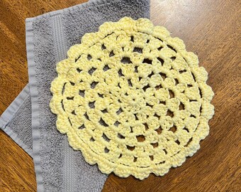 May include: A yellow crocheted doily with a delicate floral pattern, resting on a gray towel.