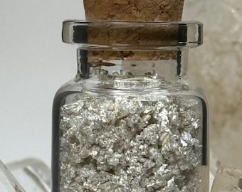 May include: A small, clear glass bottle filled with silver flakes, sealed with a cork stopper. The bottle has a rectangular shape and a narrow neck. The silver flakes appear to be metallic and reflective, filling the majority of the bottle.