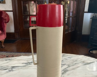 May include: A red and cream-colored thermos with a handle. The thermos has a red lid and a cream-colored body with horizontal lines.