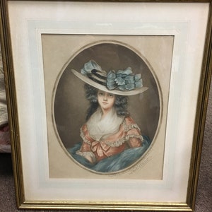 May include: A framed portrait of a woman wearing a large white hat with blue ribbons. The woman is wearing a pink dress with a lace collar and a blue shawl. The portrait is on a cream-colored background.