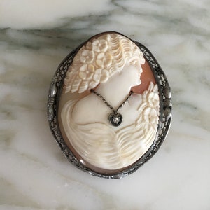 May include: Oval cameo brooch featuring a woman's profile carved in cream-colored shell. The cameo is set in a silver-toned filigree frame. A small heart-shaped pendant with a clear stone hangs from the cameo.