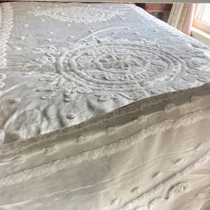 May include: A white bedspread with an intricate, raised floral and circular pattern. The bedspread has a textured, tufted design and a ruffled edge. The bedding is on a bed frame, and the overall aesthetic is soft and inviting.
