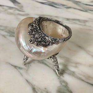 May include: A small bowl crafted from a polished iridescent seashell, featuring an ornate silver-toned metal rim with intricate floral scrollwork and three matching silver-toned metal legs that provide a stable base for the shell container.