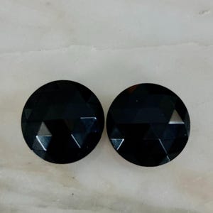 May include: Two round, faceted black gemstones. Each gem has a circular shape with multiple flat surfaces, creating a sparkling effect. The gems are displayed on a light-colored surface.