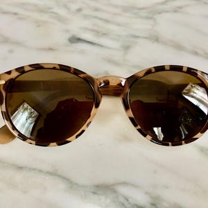 May include: Round sunglasses with a tortoiseshell pattern frame and dark lenses. The sunglasses have a classic design with a neutral color palette, suitable for various styles. The frame features a speckled pattern in shades of brown and tan.