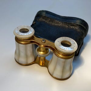 May include: Antique opera glasses with a gold-tone frame, white and gold accents, and a black leather case. The opera glasses have a pearlescent finish and are marked "PARIS". A vintage collectible.