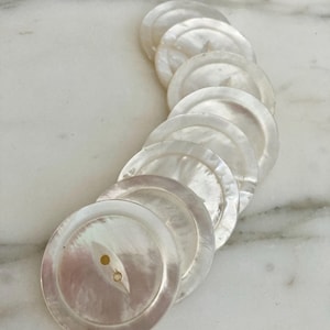 May include: A collection of iridescent, round mother-of-pearl buttons. Each button has a subtle sheen and a pair of small holes for sewing. The buttons are arranged in a curved line, suggesting a craft or sewing project.