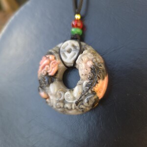 May include: A carved stone pendant necklace with a circular design. The pendant features intricate carvings in shades of gray, orange, and black. The necklace has a black cord with red, green, and gold beads.