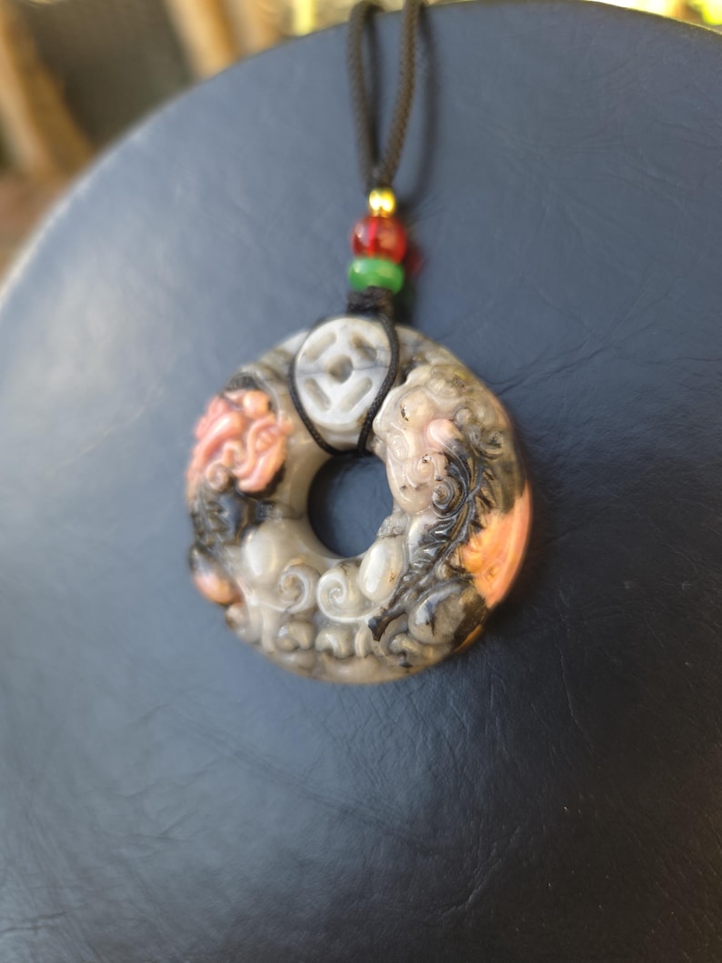 May include: A carved stone pendant necklace with a circular design. The pendant features intricate carvings in shades of gray, orange, and black. The necklace has a black cord with red, green, and gold beads.