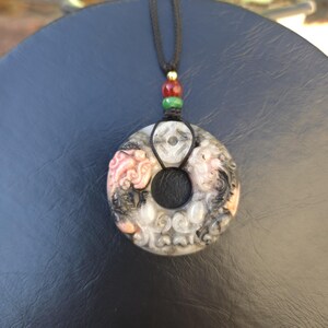 May include: A circular pendant necklace with a black cord. The pendant is gray, black, and pink with carved details and a central hole. The cord has red, green, and gold beads.