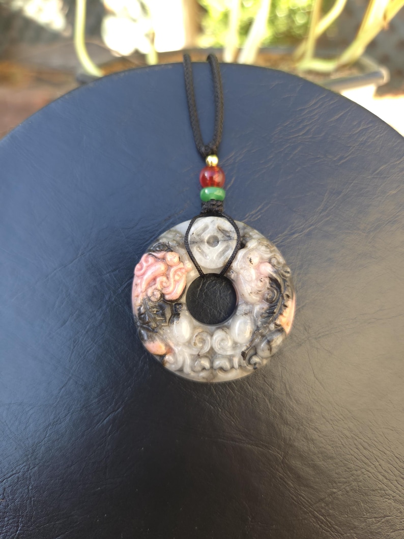 May include: A circular pendant necklace with a black cord. The pendant is gray, black, and pink with carved details and a central hole. The cord has red, green, and gold beads.