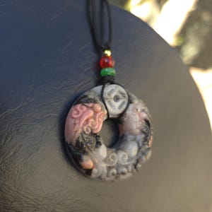 May include: A carved pendant necklace with a black cord. The pendant is circular with a detailed design in shades of pink, gray, and black. The cord has red, green, and gold beads.