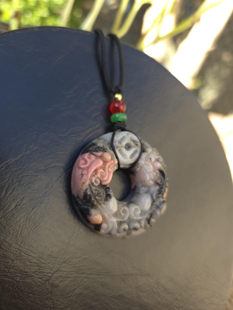 May include: A carved pendant necklace with a black cord. The pendant is circular with a detailed design in shades of pink, gray, and black. The cord has red, green, and gold beads.