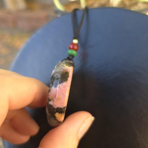 May include: A pendant necklace featuring a polished, oval-shaped stone with pink, black, and yellow patterns. The stone is suspended from a black cord with red, green, and gold beads. The necklace is held in a hand.