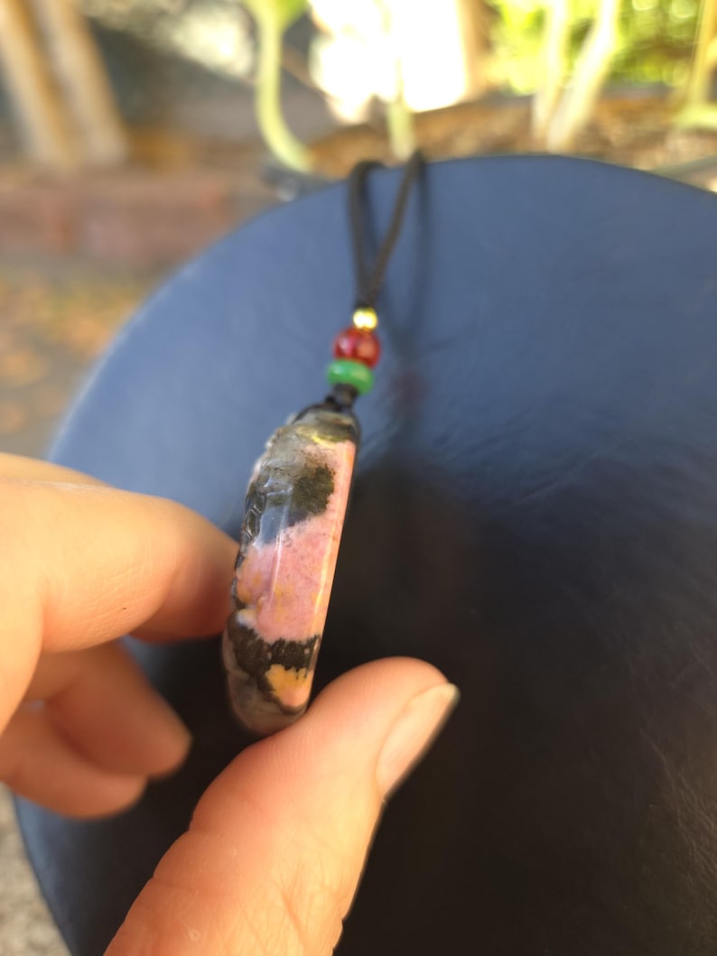 May include: A pendant necklace featuring a polished, oval-shaped stone with pink, black, and yellow patterns. The stone is suspended from a black cord with red, green, and gold beads. The necklace is held in a hand.