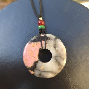 May include: A circular pendant necklace with a black cord. The pendant is a donut shape, featuring pink, black, and white marbled stone. The cord has red, green, and gold beads.