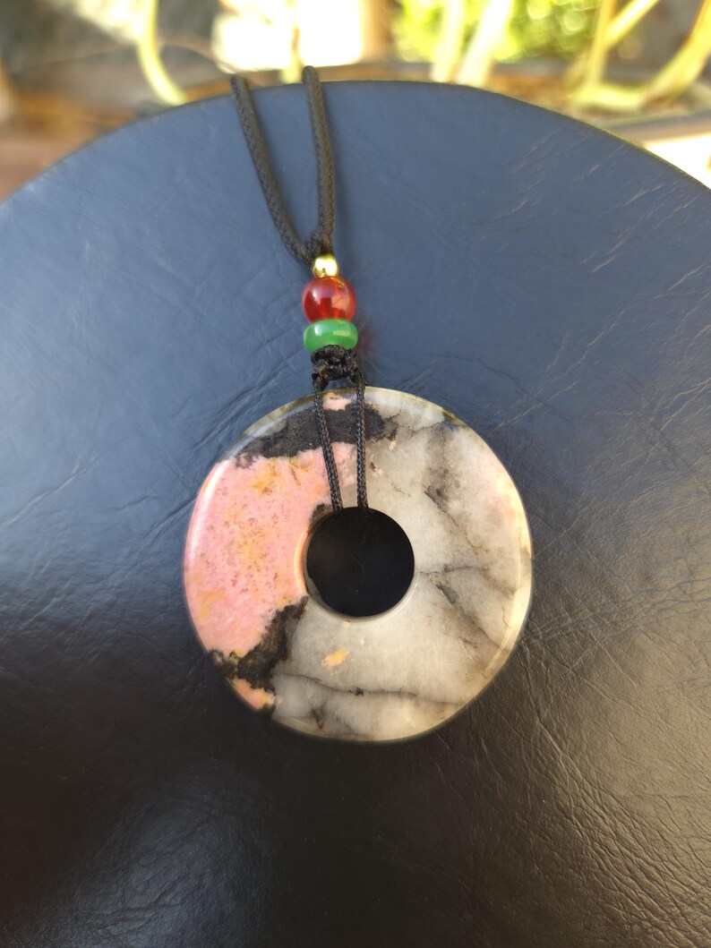 May include: A circular pendant necklace with a black cord. The pendant is a donut shape, featuring pink, black, and white marbled stone. The cord has red, green, and gold beads.
