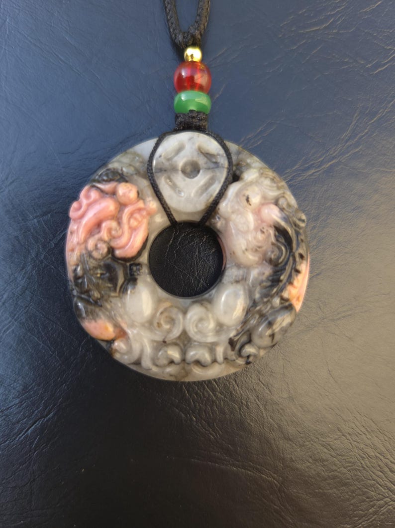 May include: A carved pendant with a circular design, featuring intricate relief carvings in shades of gray, pink, and black. The pendant is suspended from a black cord with red, green, and gold beads. The pendant has a central hole and a black leather backing.
