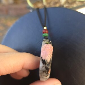 May include: A pendant necklace featuring a rectangular stone with pink, black, and white patterns. The stone is suspended from a black cord with red, green, and gold beads. The pendant is held between fingers.