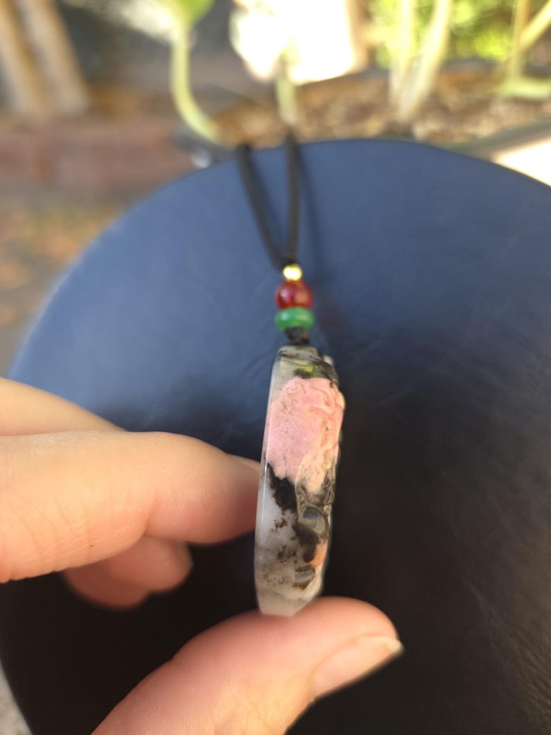 May include: A pendant necklace featuring a rectangular stone with pink, black, and white patterns. The stone is suspended from a black cord with red, green, and gold beads. The pendant is held between fingers.