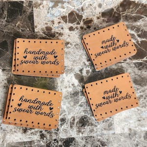 May include: Four rectangular, tan leather tags with black text that reads "handmade with swear words" and "made with swear words". Each tag has a border of small, evenly spaced holes and a heart symbol.