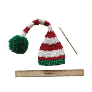 May include: A handmade crochet elf hat with red and white stripes, a green pom-pom, and a long, curved tip. A metal crochet hook and a wooden ruler are also in the image. The ruler measures up to 12 inches.