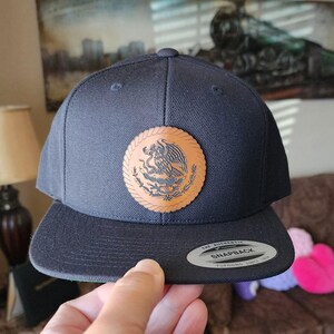 May include: A navy blue snapback hat with a brown leather patch featuring the Mexican coat of arms. The hat has a flat brim and a snap closure. The text "THE AUTHENTIC SNAPBACK YUPOONG SINCE 1974" is visible on the brim.