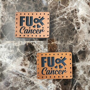 May include: Two rectangular, tan leather patches with the words "FU Cancer" cut out in black. The design includes a ribbon, hearts, and small dotted edges. The patches are displayed on a brown marble surface.