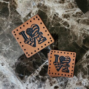 May include: Two square tan leather patches featuring a dark engraved design of the word LOVE with a heart shaped like an American flag replacing the letter O. Each patch has a series of small pre-punched holes along all four edges for sewing.