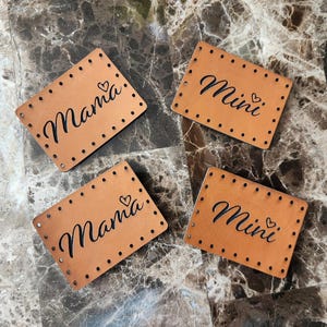 May include: Four rectangular, light brown leather patches. Two patches read "Mama" with a heart, and two read "Mini" with a heart, all in black script. Each patch has evenly spaced holes around the edges.