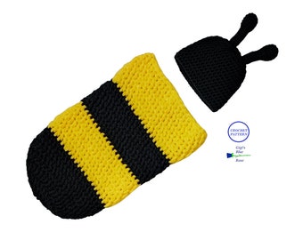 May include: A handmade crochet baby cocoon and hat set in a bee design. The cocoon is yellow and black striped, and the hat is black with two antennae. The set is designed for newborn photography props.